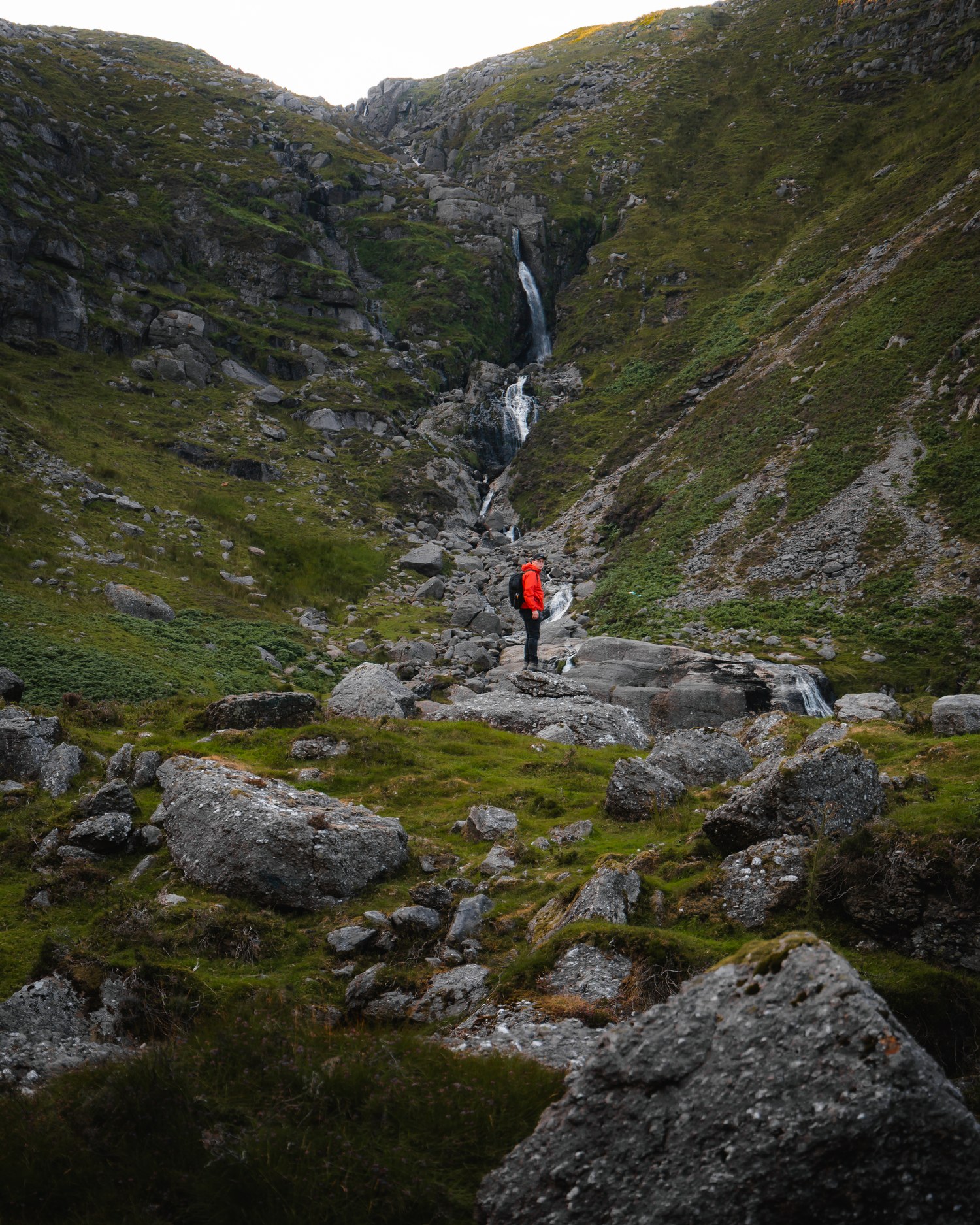 Walker in a red jacket below a mountain waterfall