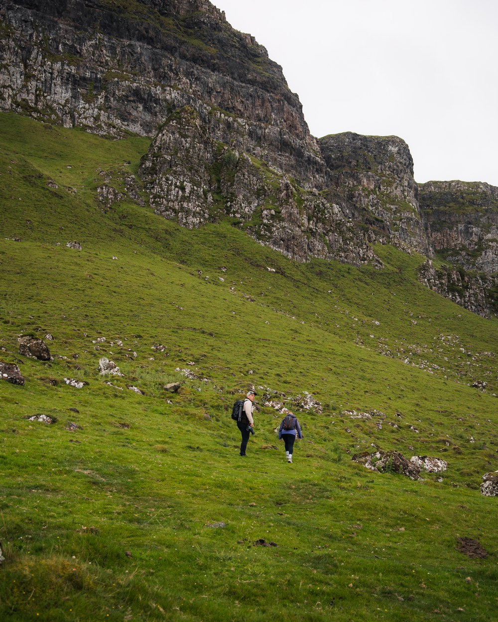Two walkers crossing a green slope below a rocky escarpment
