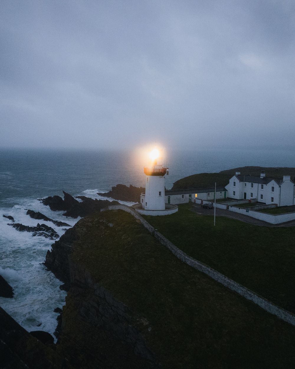Irish lighthouse with its beam lit at blue hour above stormy cliffs