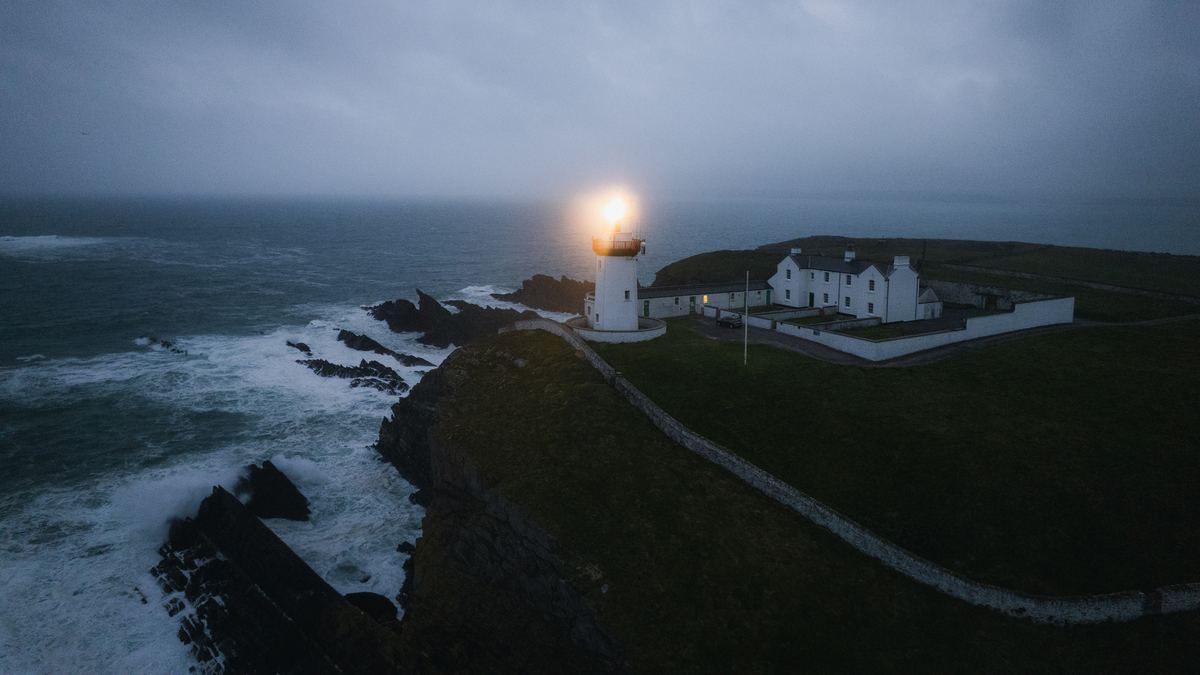 Irish lighthouse with its beam lit at blue hour above stormy cliffs
