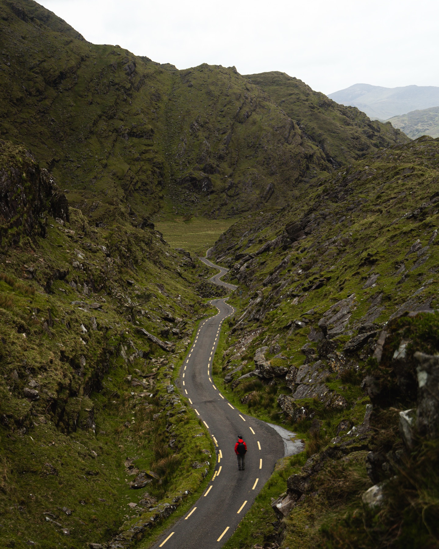 A hiker in a red jacket walking the road through Ballaghbeama Gap between steep rock walls, County Kerry